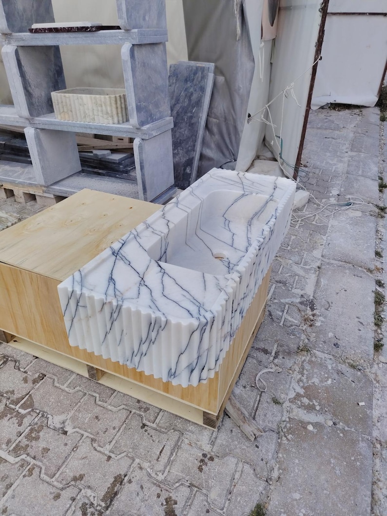 May include: A rectangular white marble sink featuring prominent dark blue veining and a fluted, vertically ribbed exterior design. The basin has a smooth interior surface with a central drain hole, resting on a light-colored wooden support structure.
