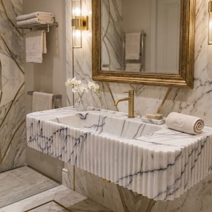 May include: A wall-mounted white marble sink featuring a fluted, ribbed exterior design with dark grey veining. The sink includes a gold-toned faucet, a matching soap dish, and a rolled white towel resting on the flat surface of the basin.