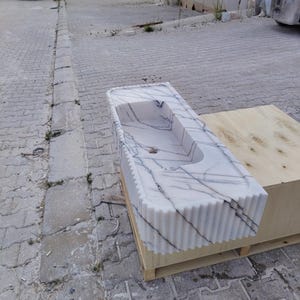 May include: A rectangular white marble sink featuring prominent dark grey veining and a fluted, vertically ribbed exterior design. The basin has a smooth interior surface with a central drain hole, showcasing a polished stone finish throughout.