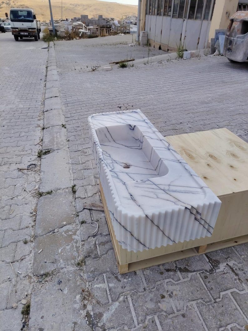 May include: A rectangular white marble sink featuring prominent dark grey veining and a fluted, vertically ribbed exterior design. The basin has a smooth interior surface with a central drain hole, showcasing a polished stone finish throughout.