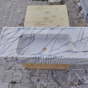 May include: A rectangular white marble sink featuring prominent dark blue veining throughout the surface. The front edge of the sink has a vertical fluted or ribbed texture, and the basin includes a centered circular drain hole opening.