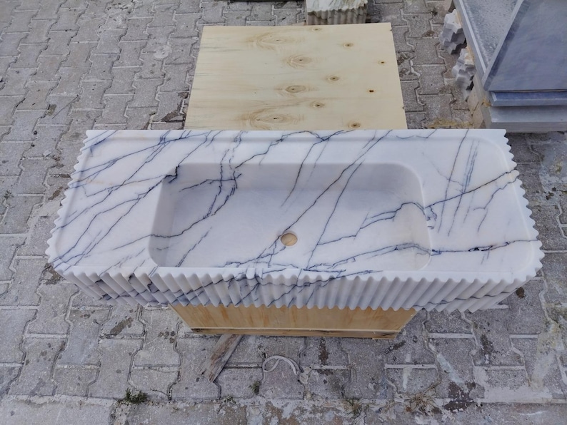 May include: A rectangular white marble sink featuring prominent dark blue veining throughout the surface. The front edge of the sink has a vertical fluted or ribbed texture, and the basin includes a centered circular drain hole opening.