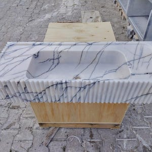 May include: A rectangular white marble sink featuring prominent dark blue veining throughout the stone. The exterior front and side edges of the basin are designed with a vertical fluted texture, and a central drain hole is visible at the base.