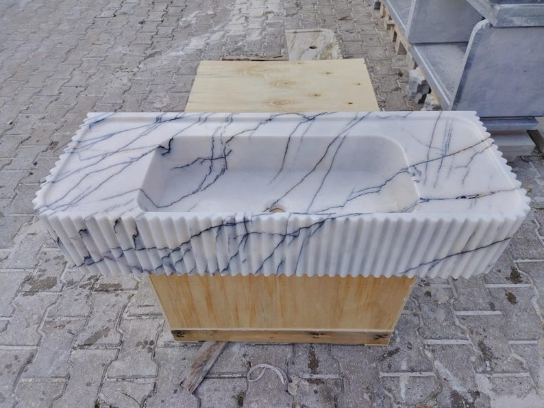 May include: A rectangular white marble sink featuring prominent dark blue veining throughout the stone. The exterior front and side edges of the basin are designed with a vertical fluted texture, and a central drain hole is visible at the base.