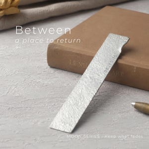 Peut inclure: Un marque-page rectangulaire en métal argenté avec une surface texturée et la date 06.10.16 estampée. Le marque-page repose contre un livre marron, avec le texte Between a place to return et MOON SERIES - Keep what fades visible.