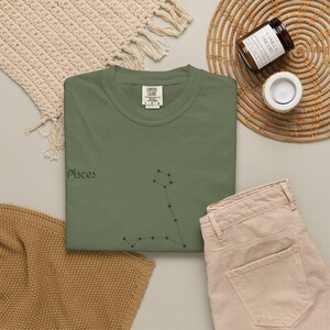 An olive green t-shirt with "Pisces" and the Pisces constellation in black, laid flat next to beige pants and a candle.