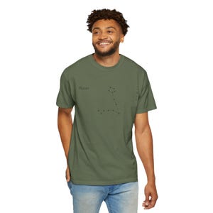 A man in an olive green t-shirt with "Pisces" and the Pisces constellation printed in black, standing with one hand in his pocket.
