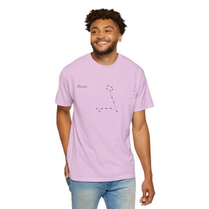A man in a light purple t-shirt with "Pisces" and the Pisces constellation printed in black, standing with one hand in his pocket.
