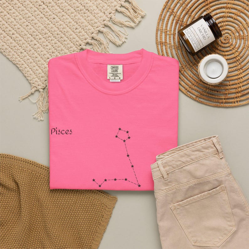 A hot pink t-shirt with "Pisces" and the Pisces constellation in black, laid flat next to beige pants and a candle.