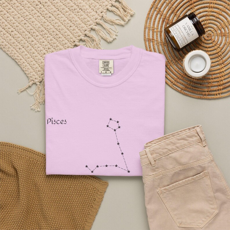 A light pink t-shirt with "Pisces" and the Pisces constellation in black, laid flat next to beige pants and a candle.