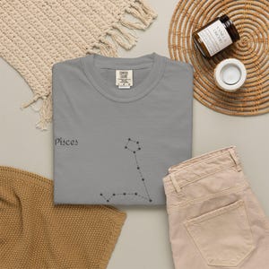 A grey t-shirt with "Pisces" and the Pisces constellation in black, laid flat next to beige pants and a candle.