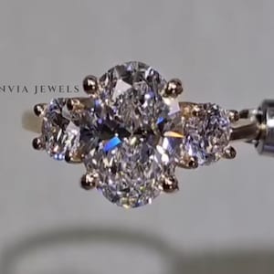 May include: A three-stone ring featuring a central oval-cut diamond flanked by two smaller round-cut diamonds, all set in a gold band. The text ZENVIA JEWELS is visible on the left and bottom right. The ring is held by a metal tool.