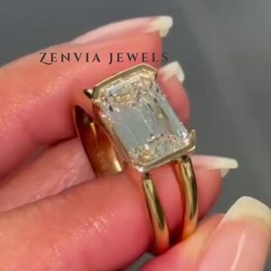 May include: A gold ring featuring a large, rectangular emerald-cut clear gemstone set in a bezel setting. The band is split into two thin, parallel gold wires that connect to the base of the stone setting. The text ZENVIA JEWELS is visible.