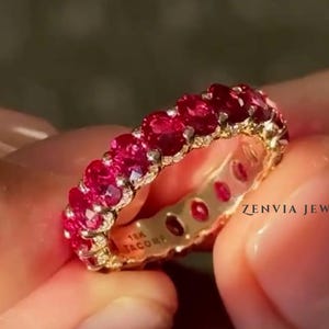 以下が含まれることがあります： 18金ゴールドのエタニティリングで、鮮やかなオーバルカットの赤いルビーが一列に並んでいます。リングの内側には「18K TACORI」および「ZENVIA JEWELS」の刻印があります。磨き上げられたゴールドのセッティングが、それぞれの宝石をしっかりと固定しています。