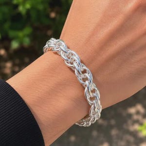 May include: A polished silver chain bracelet featuring a series of interlocking, rounded links that create a thick, textured, and continuous circular band. The metal surface reflects light, highlighting the intricate, repeating pattern of the links.