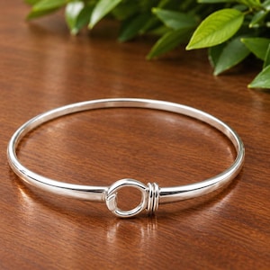 May include: A polished silver bangle bracelet featuring a circular hook-and-eye style clasp mechanism at the center. The bracelet has a smooth, rounded band that tapers slightly toward the front where the two ends meet in a secure loop closure.