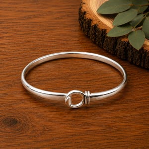 Könnte beinhalten: Ein polierter Silberarmreif mit einem kreisförmigen Hakenverschluss in der Mitte. Das Armband hat ein glattes, abgerundetes Band, das sich zur Vorderseite hin leicht verjüngt, wo der kreisförmige Verschluss durch drei kleine, parallele Silberringe gesichert ist.