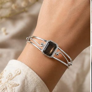 May include: A silver cuff bracelet featuring a central rectangular faceted smoky quartz gemstone. The metal band consists of two parallel wires that curve into decorative scrolls on either side of the stone setting, creating an open design.
