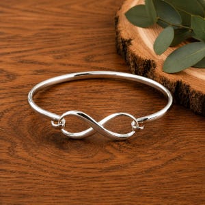 May include: A polished silver bangle bracelet featuring a central infinity symbol design. The circular band is crafted from smooth, reflective metal, with the infinity loop securely integrated into the front, creating a continuous, sleek, and metallic aesthetic.