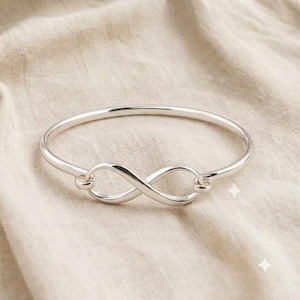 Puede incluir: Una pulsera rígida de plata pulida que presenta un símbolo de infinito en el centro. El metal tiene una superficie lisa y reflectante con una banda circular continua que se conecta al bucle del infinito, creando un diseño sencillo.