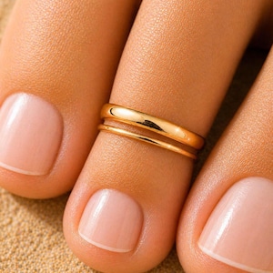 Puede incluir: Un anillo para el dedo del pie en tono dorado pulido con un diseño de doble banda, que presenta una banda redondeada más gruesa y una banda más fina y lisa superpuestas. El metal tiene un acabado brillante y reflectante que capta la luz.