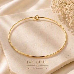 May include: A thin, circular 14K gold bangle bracelet featuring a simple hook-and-eye closure mechanism. The jewellery piece is polished to a high shine, reflecting light along its smooth, rounded metal surface in a continuous, elegant loop.