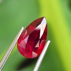 May include: A pear-shaped, faceted red gemstone held by silver-toned metal tweezers. The stone features a deep crimson hue with internal clarity and light-reflecting facets across its surface, showcasing a pointed top and a rounded base.