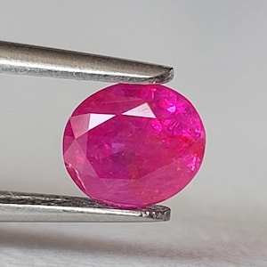 Op de afbeelding: Een levendige, ovaal geslepen edelsteen met een diepe magenta-roze tint en een gefacetteerd oppervlak. De steen wordt stevig vastgehouden door een zilverkleurige metalen pincet, wat de doorschijnende kwaliteit en lichtreflecties toont.