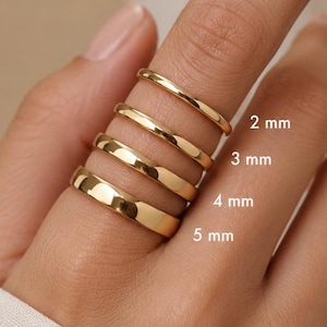 May include: Four polished gold-tone bands stacked on a finger, featuring varying widths labelled as 2 mm, 3 mm, 4 mm, and 5 mm. Each ring has a smooth, rounded profile and a reflective metallic finish, showcasing the different band thicknesses clearly.