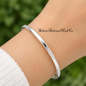Puede incluir: Una pulsera rígida de plata pulida con una superficie lisa y redondeada. El metal tiene un acabado reflectante y un diseño circular simple y continuo. El texto SilverArtisanHubCo es visible en letras cursivas de color negro.