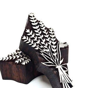 May include: Two dark brown wooden printing blocks featuring a carved wheat sheaf design. The raised surfaces of the wheat stalks and the tied base are highlighted in white, creating a high-contrast pattern for textile or paper stamping.