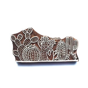 Könnte beinhalten: Ein brauner Holzstempel mit einem geschnitzten Design aus verschiedenen Kakteen und Sukkulenten. Die erhabenen weißen Oberflächen zeigen einen Feigenkaktus, einen runden Kugelkaktus, einen hohen Säulenkaktus und weitere Wüstenpflanzen.