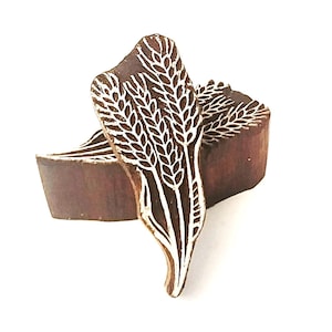 May include: A dark brown wooden printing block featuring a carved design of three wheat stalks. The raised surface of the wheat pattern is light in color, contrasting with the dark wood grain of the block. The block has a thick, solid construction.