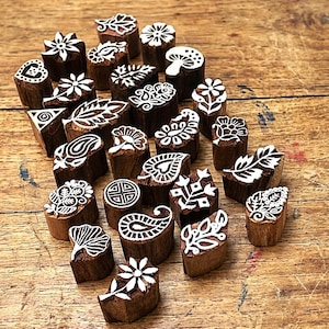 Puede incluir: Una colección de pequeños bloques de madera cilíndricos con diseños blancos tallados en sus superficies planas. Las tallas incluyen varios motivos botánicos como flores, hojas, patrones de cachemira y un hongo, dispuestos en un grupo disperso.