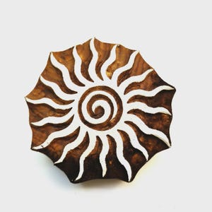 May include: A brown wooden printing block featuring a carved sun design with a central spiral and radiating wavy rays. The raised surface of the sun motif is white, contrasting with the natural dark brown wood grain of the circular block.