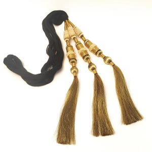 May include: A decorative tassel accessory featuring three long, golden-brown silk-like fringe strands attached to a black cord. Each strand is adorned with cylindrical gold-toned metal beads and small, cream-colored spherical bead accents.