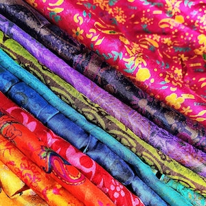 Puede incluir: Una colección de tiras de tela dobladas dispuestas en una pila diagonal. Los textiles presentan colores vibrantes que incluyen rosa intenso, morado oscuro, verde lima, azul brillante, rojo, naranja y amarillo, todos decorados con intrincados patrones florales y de cachemira.