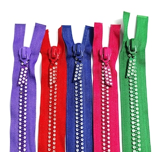 Puede incluir: Cinco cremalleras de tela largas en colores morado, rojo, azul, rosa y verde. Cada cremallera cuenta con un tirador a juego y una tira decorativa de brillantes transparentes a lo largo de los dientes de la cremallera.
