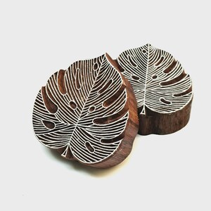 May include: Two dark brown wooden printing blocks, each carved into the shape of a monstera leaf. The surface of each block features intricate, raised white lines that form the detailed vein pattern of the leaf, designed for textile printing.
