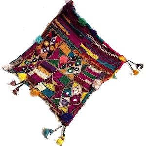 May include: A square textile wall hanging featuring a deep magenta base with intricate multi-colored embroidery in yellow, green, and blue. The piece includes geometric patterns, small circular mirrors, and decorative tassels on the corners.