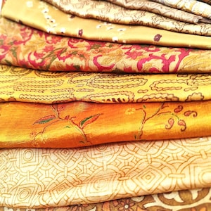 May include: A stack of folded fabric pieces in various shades of gold, orange, and brown. Each layer features distinct patterns including floral motifs, geometric shapes, paisley designs, and intricate line work, all displayed in a layered arrangement.