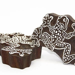 May include: Two dark brown wooden printing blocks featuring a carved frog design. The raised surfaces of the frog motifs are highlighted in white, showcasing detailed webbed feet and a textured body pattern on each individual block.