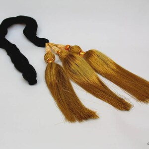 May include: A decorative accessory featuring a thick, braided black cord attached to three long, metallic gold tassels. Each tassel is secured with a small, textured gold-toned bead and a section of tiny, cream-colored beads at the top attachment point.