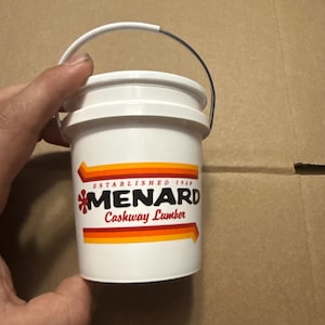 May include: A small white plastic bucket with a metal handle. The front features a logo with orange and red stripes, the text "ESTABLISHED 1960", the word "MENARD" in bold black letters, and the text "Cashway Lumber" in red script lettering.