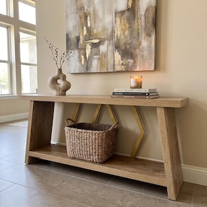 May include: A light oak console table with a lower shelf and gold-toned geometric metal supports. On the table sits a beige abstract ceramic vase with branches, a lit candle, and two stacked books. A woven seagrass basket rests on the lower shelf.