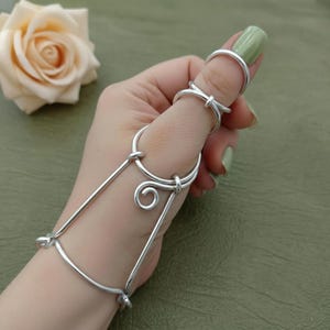 Può includere: Una catena da mano argentata con più anelli e barre di collegamento. Il design include un anello per il pollice, un anello con un nodo e una struttura simile a un braccialetto collegata da barre dritte e un ricciolo decorativo.