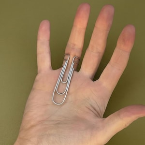 May include: A silver paperclip bent into a ring shape, worn on a finger. The paperclip is a simple, everyday office supply, repurposed as a piece of quirky jewelry. The background is a solid olive green.