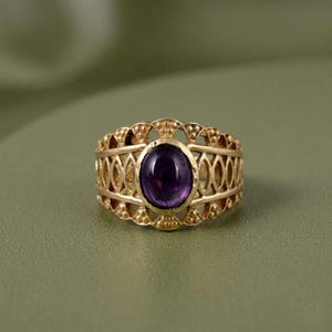 May include: A gold-toned ring featuring an oval-shaped, deep purple cabochon gemstone set in the centre. The band is crafted with an intricate, openwork lattice design that includes small, raised bead-like details along the top and bottom edges.