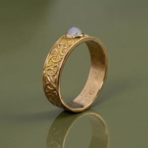 Op de afbeelding: Een goudkleurige ring met een Keltisch knoopontwerp en een kleine, lichtgekleurde steen. De ring heeft een brede band met gedetailleerde reliëfpatronen. De steen is bovenop de ring gezet.