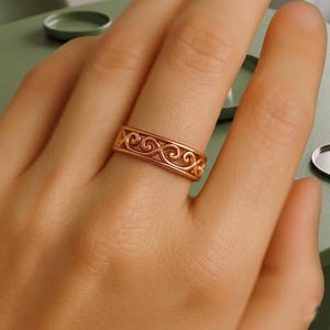 May include: A rose gold ring with an openwork design featuring swirling patterns. The ring is worn on a finger, showcasing its intricate details and warm metallic hue. The ring's design is symmetrical and elegant.